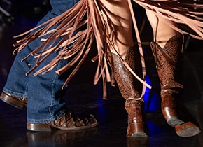 A close-up of the legs of two people in cowboy boots dancing on a stage.